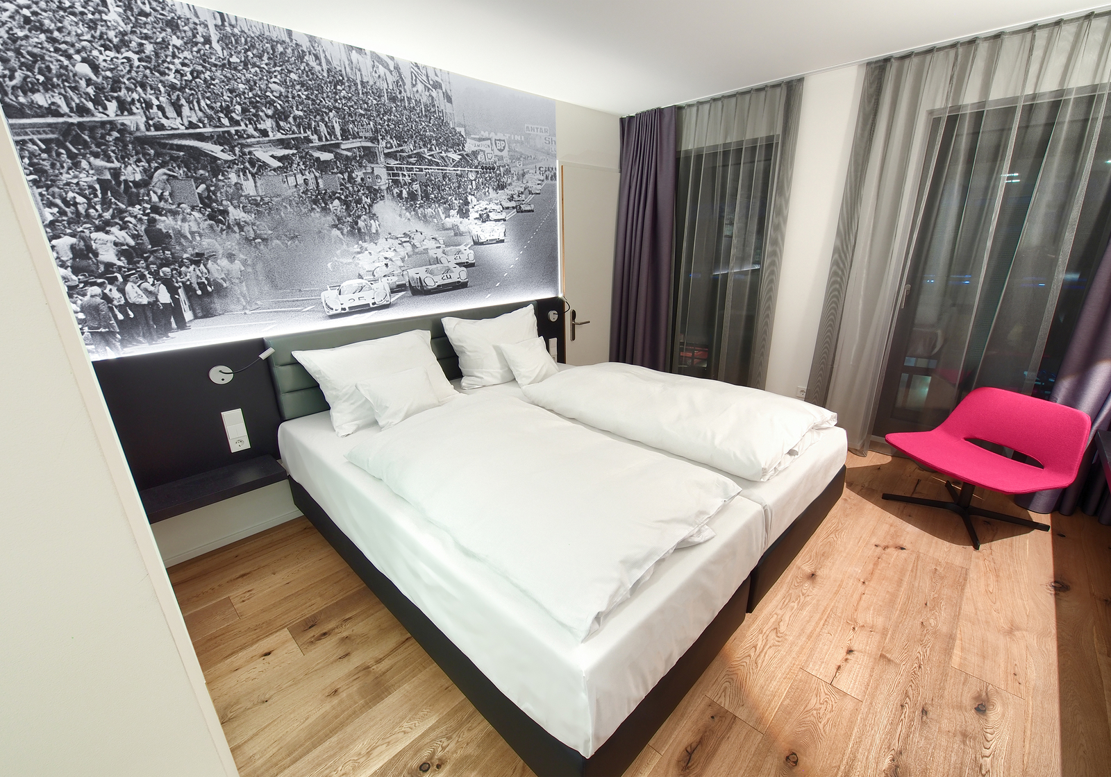 Modernes Doppelzimmer mit einem gemütlichem Bett neben einem pinken Sessel und bodentiefen Fenstern.