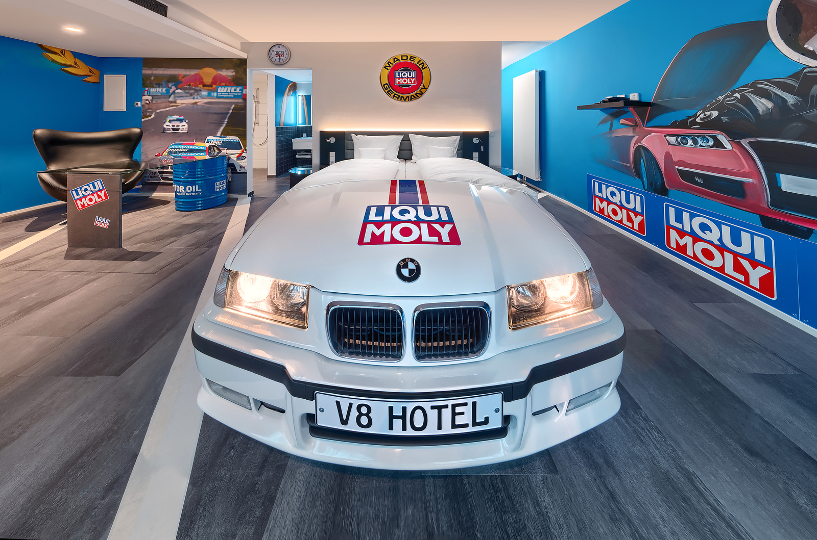 Weißes BMW Autobett im Autozimmer Liqui Moly im V8 Hotel neben einer blauen Wand mit Liqui Moly Logos.