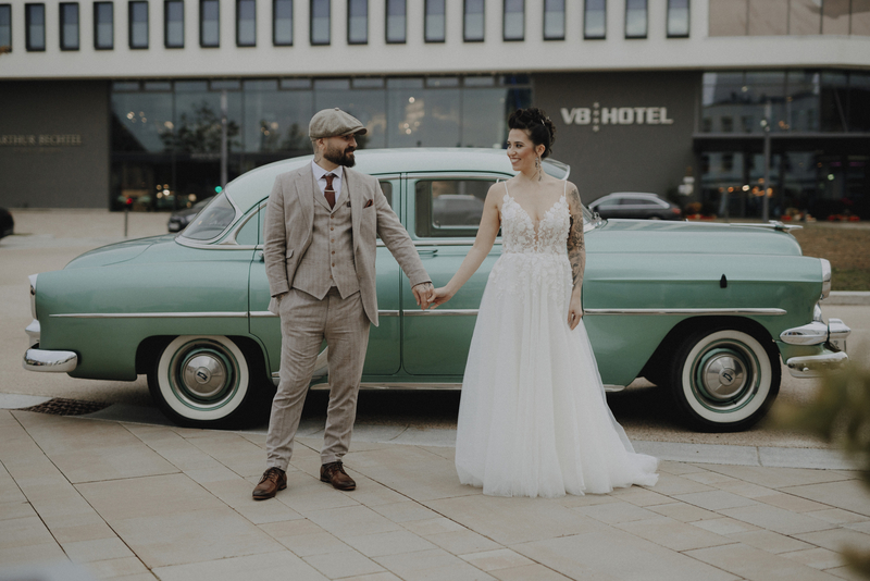 Eine Frau in Hochzeitskleid hält lächelnd die Hand ihres Mannes vor einem grünen Auto, im Hintergrund das V8 Hotel.
