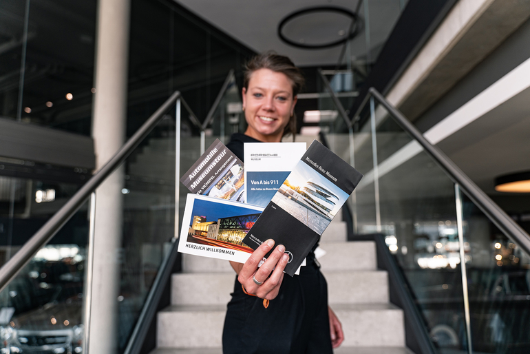 Eine junge Frau hält lächelnd vier Broschüren mit Sehenswürdigkeiten rund um Stuttgart in die Kamera.
