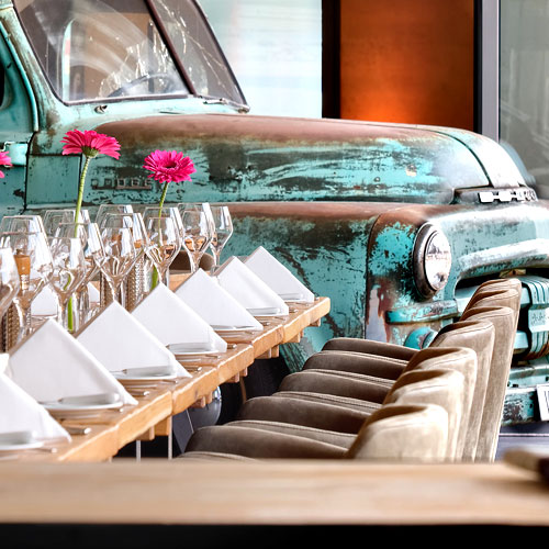 Ansprechend eingedeckter Tisch mit farbigen Blumen, Weingläsern und einem türkisen Oldtimer im Hintergrund des PICK-UP Restaurants.