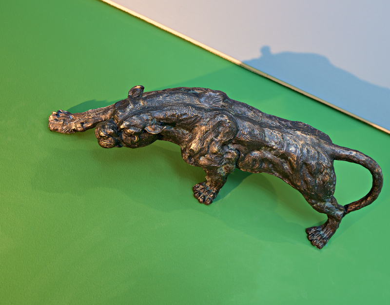 Eine bronzene Skulptur eines Jaguars, das sich vorwärts streckt, mit detaillierten Texturen auf dem Körper. Die Statue befindet sich auf einem grünen Hintergrund, der die Konturen des Tieres betont.