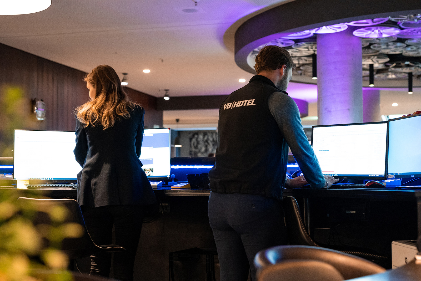 Zwei Mitarbeiter stehen an Arbeitsplätzen in einem Hotel. Jeder arbeitet an einem Computer mit mehreren Bildschirmen. Die Atmosphäre ist modern und leicht beleuchtet, mit einem geschäftigen, professionellen Umfeld.