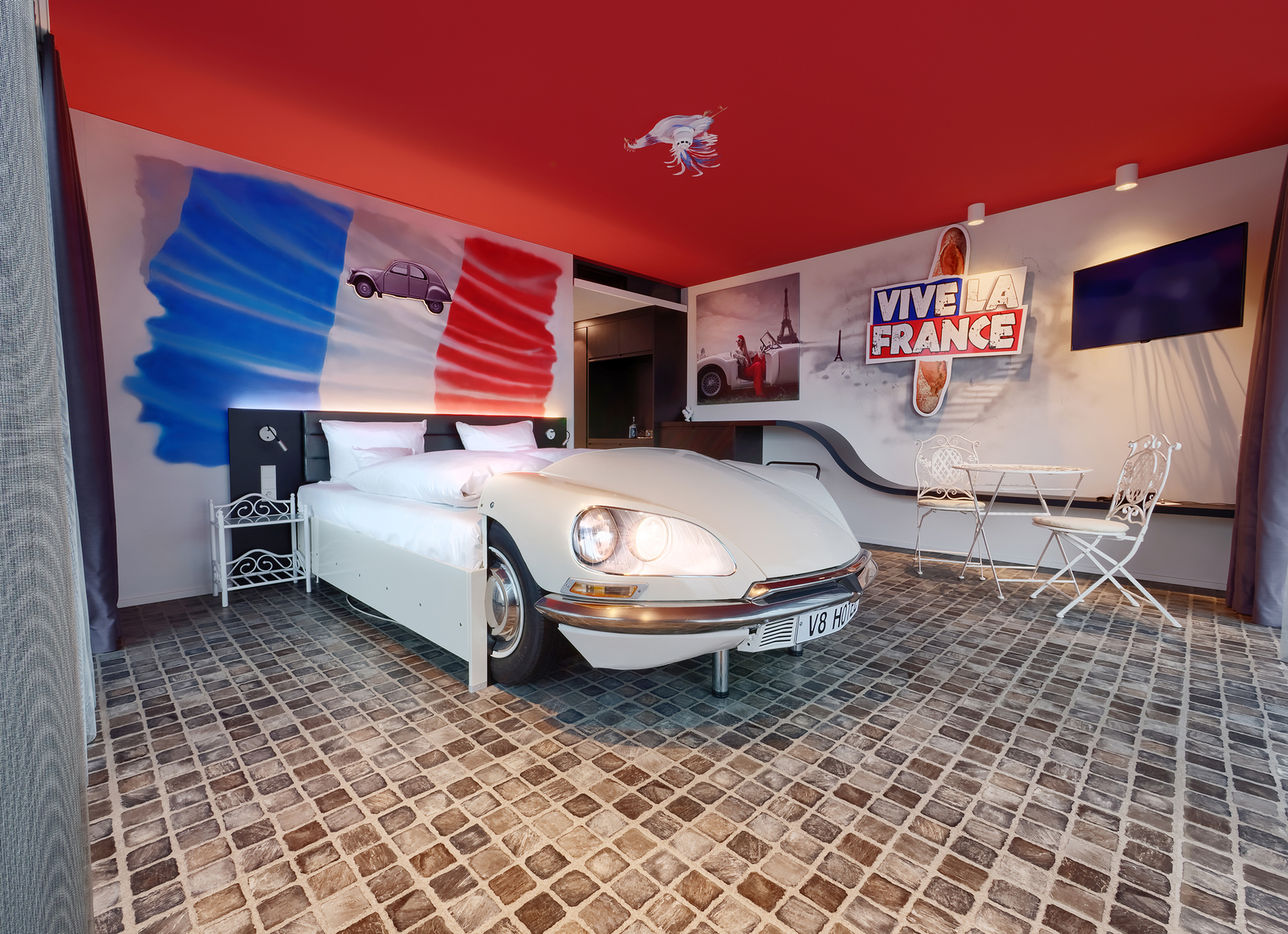 Frankreich-Themenzimmer im V8 Hotel eingerichtet mit zwei Metallstühlen an einem weißen Metalltisch, einer Frankreichflagge über dem Autobett und einem Baguette Schild an der Wand