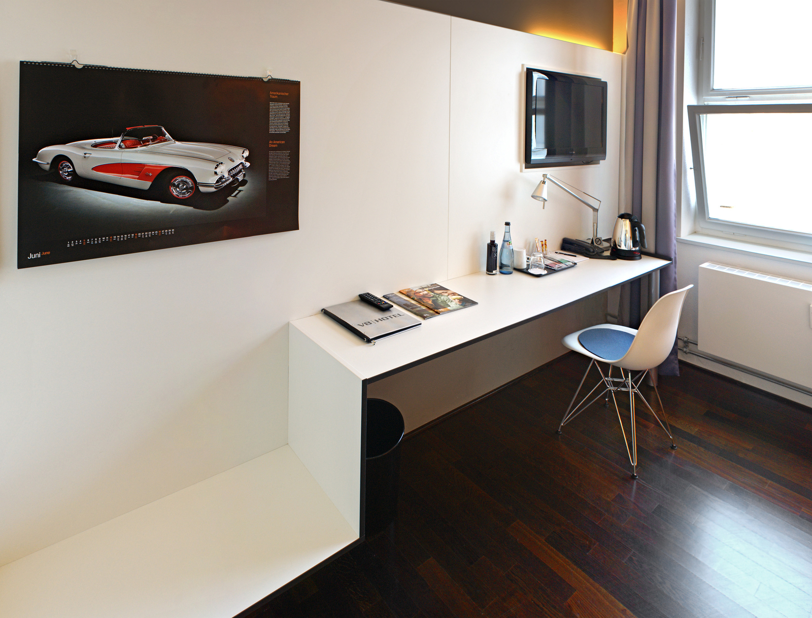 Ein modernes Hotelzimmer mit einem Schreibtisch, einem Fernseher an der Wand, einem Kalender mit einem Oldtimer, einer Kaffeemaschine und Wasserflaschen. Der Boden ist aus dunklem Holz, und ein Stuhl mit einem blauen Sitzpolster steht am Tisch.