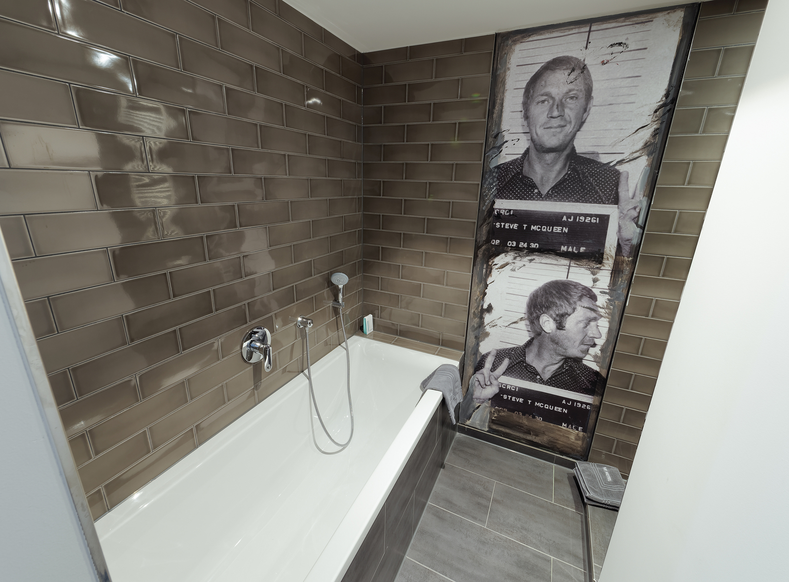 Das Bild zeigt ein modernes Badezimmer mit einer Badewanne und einer gefliesten Wand in braunen Tönen. An einer Wand ist ein großes, historisches Foto einer Person mit einem Polizeibild zu sehen, das an Steve McQueen erinnert.