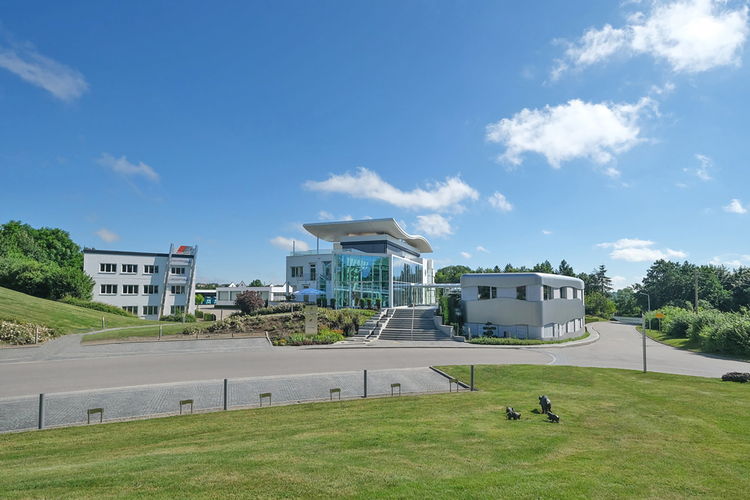 Ein modernes Gebäude mit Glasfront und einem markanten Dach steht im Vordergrund. Im Hintergrund sind weitere Gebäude und eine grüne Landschaft sichtbar. Der Himmel ist blau mit einigen Wolken, und eine steinerne Auffahrt führt zu den Eingängen.