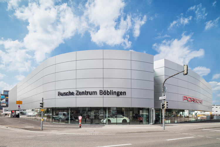 Das Bild zeigt das Porsche Zentrum Böblingen, ein modernes, großes Gebäude mit einer geschwungenen, silbernen Fassade. An der Vorderseite sind die Aufschriften „Porsche Zentrum Böblingen“ und das Porsche-Logo sichtbar. Im unteren Bereich sind Fenster zu sehen, durch die Fahrzeuge im Inneren erkennbar sind.