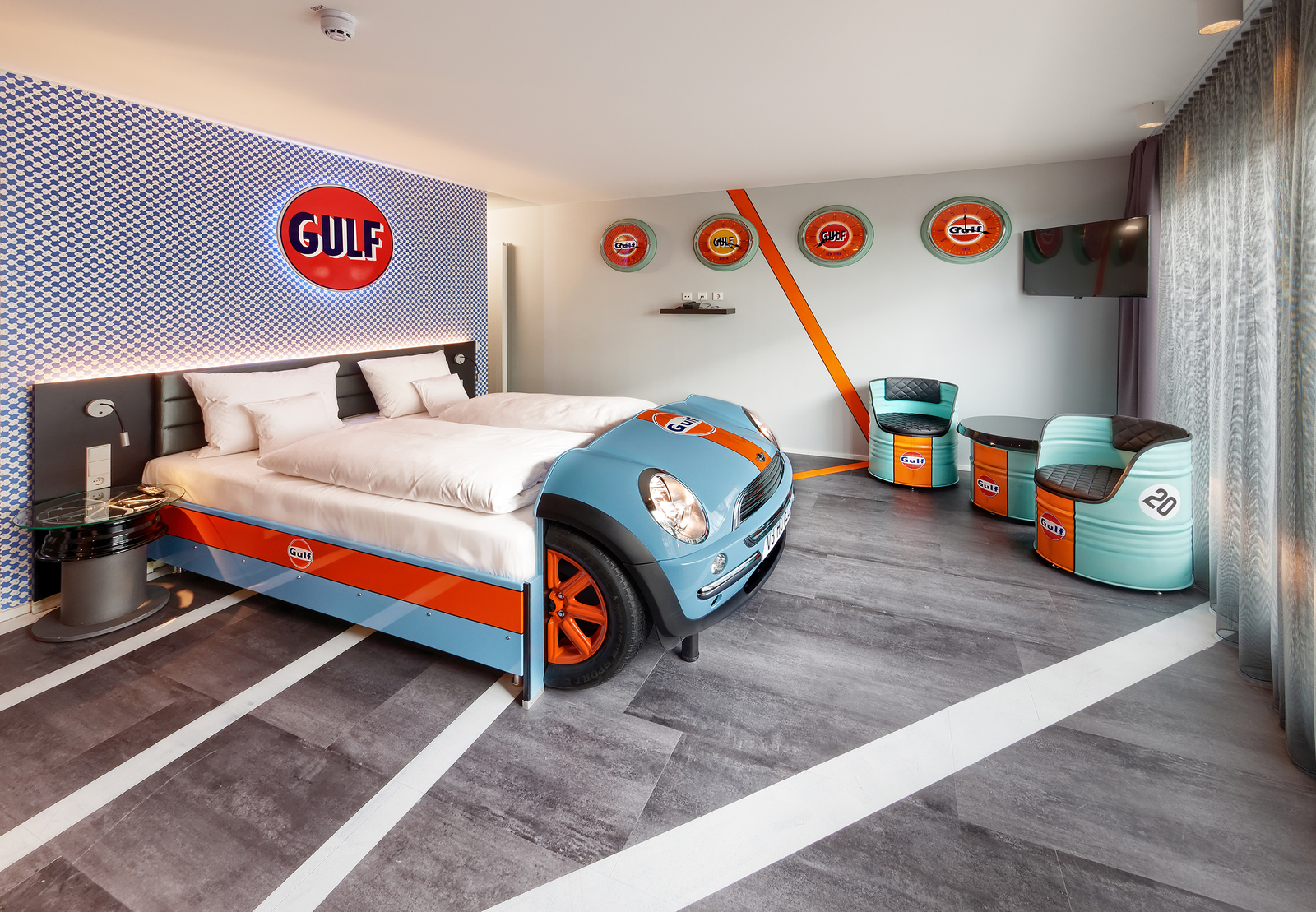 Das Bild zeigt ein automobil-inspiriertes Hotelzimmer mit einem Bett, das wie ein Rennwagen gestaltet ist. Die Wände sind blau und kariert, mit dem Logo "Gulf". Es gibt zwei stilisierte Stühle und einen Couchtisch in entspannter Anordnung. Die Einrichtung ist kreativ und thematisch gestaltet.