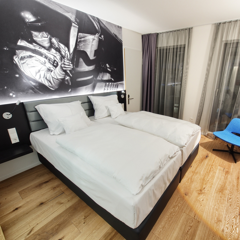 Blick auf ein gemütliches Doppelbett unter einem schwarz-weiß Foto eines Rennfahrers in seinem Auto im Themenzimmer im V8 Hotel