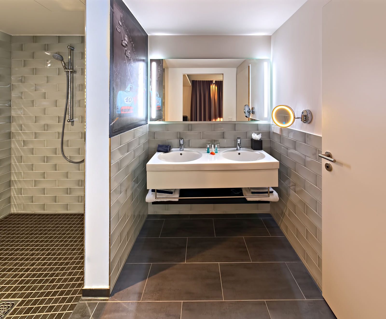Das Bild zeigt ein modernes Badezimmer mit einer Dusche und einem Waschtisch mit zwei Waschbecken. Über dem Waschbecken sind Spiegel und eine runde LED-Leuchte angebracht. Die Wände sind gefliest und der Boden ist dunkel. Das Ambiente wirkt hell und einladend.