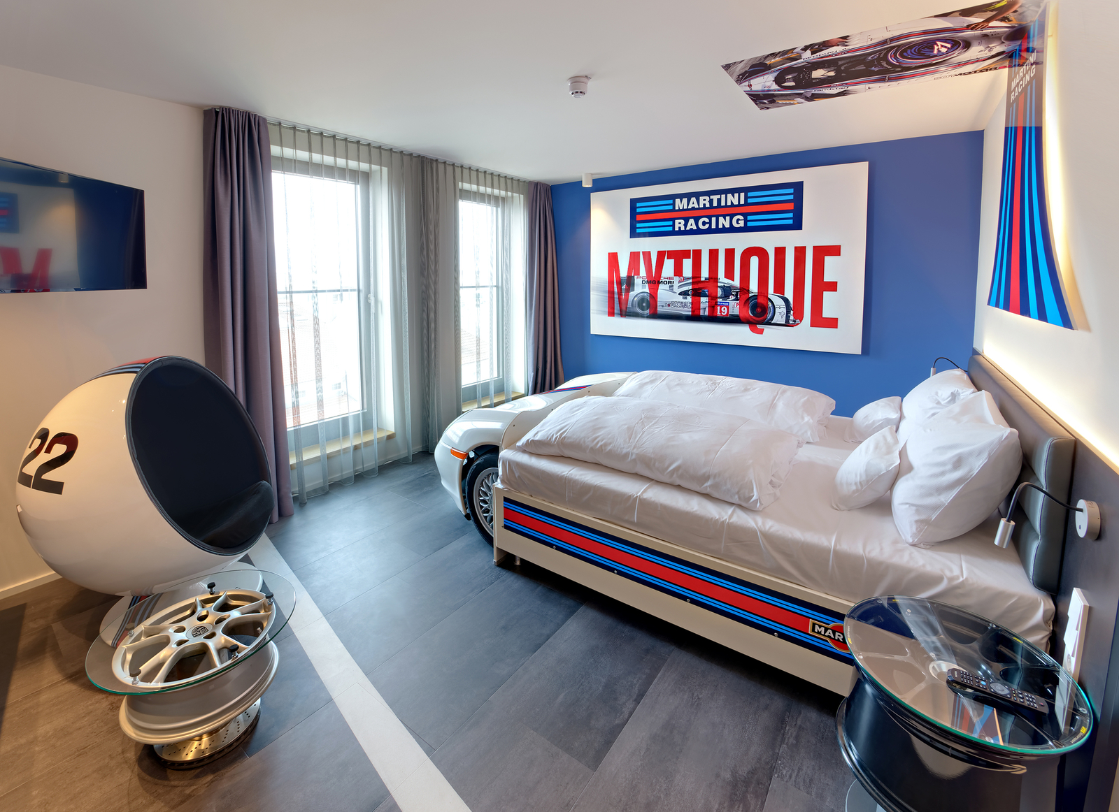 Das Bild zeigt ein modernes Zimmer mit einem Rennmotorsport-Thema. Ein Bett in Form eines Rennwagens befindet sich vor einer blauen Wand mit einem großen Martini Racing-Poster. Neben dem Bett steht ein runder Stuhl und ein Tisch in sportlichem Design. Große Fenster sorgen für Tageslicht.