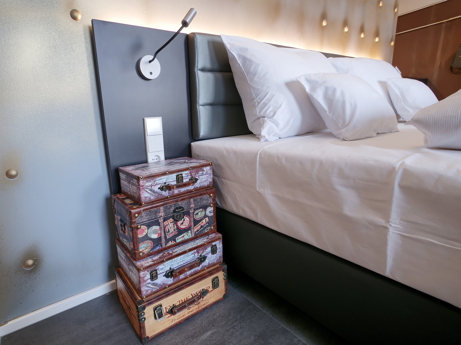 Ein modernes Schlafzimmer mit einem großen Bett, das mit weißen Kissen dekoriert ist. Neben dem Bett steht ein Stapel von drei alten Reisekoffern in unterschiedlichen Designs. An der Wand hängt ein Licht.