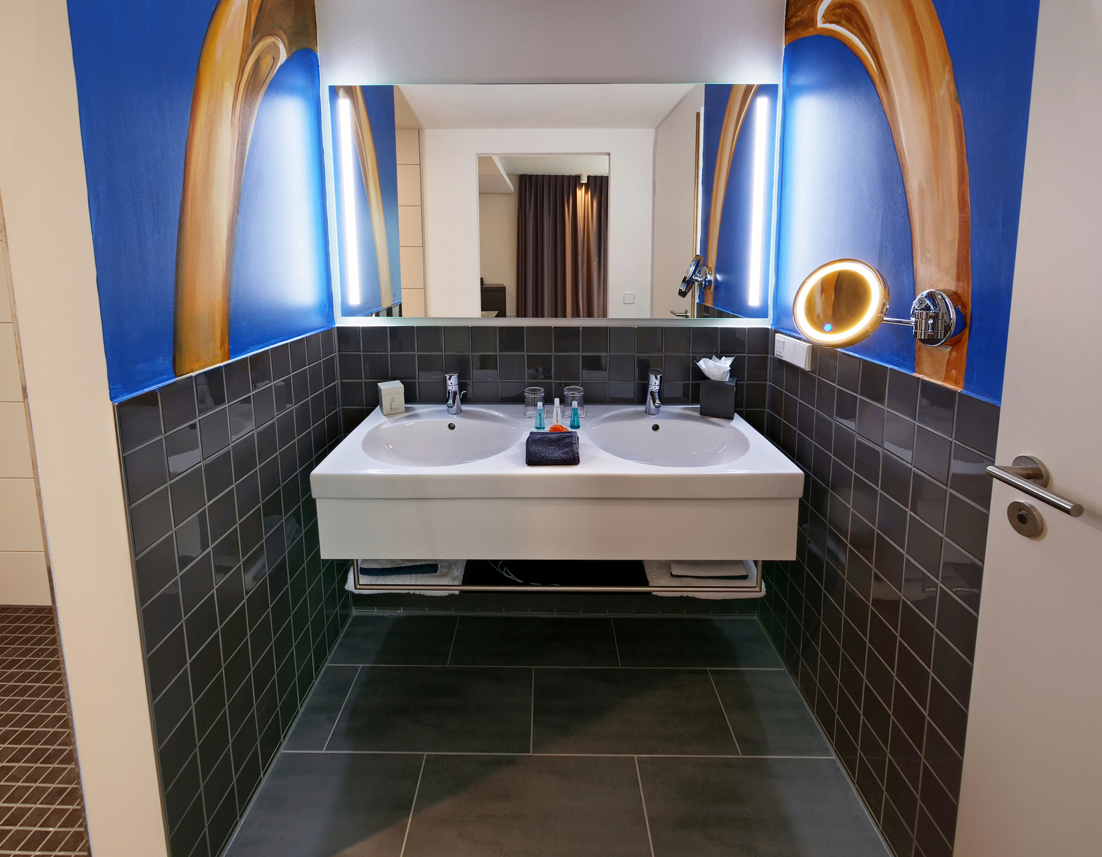 Das Bild zeigt ein modernes Badezimmer mit einem doppelten Waschbecken, das in eine graue Fliesenwand integriert ist. An der Wand befinden sich dekorative Elemente in Blau und Gold. Auf einem Handtuch liegen Pflegeprodukte in kleinen Fläschchen, und ein Spiegel ist mit einer ringförmigen Leuchte ausgestattet.