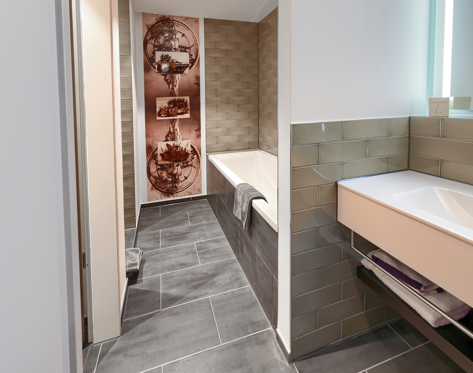 Das Bild zeigt ein modernes Badezimmer mit einer freistehenden Badewanne, hellen Fliesen und einem Waschbecken auf einer eleganten Ablage. Eine Wand mit dekorativen Elementen zieht Aufmerksamkeit auf sich. Handtücher liegen bereit, und der Boden ist mit dunklen Fliesen ausgelegt.