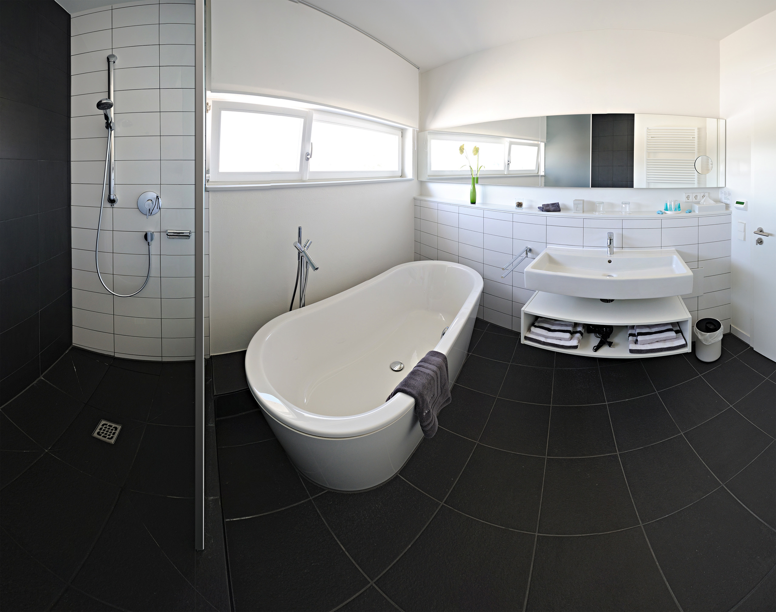 Ein modernes Badezimmer mit einer freistehenden Badewanne und einer separaten Dusche. Ein Waschbecken mit Unterbau ist vorhanden, darunter liegen Handtücher. Große Fenster sorgen für viel Tageslicht, und eine grüne Blume steht auf dem Waschtisch. Der Boden ist mit dunklen Fliesen ausgelegt.