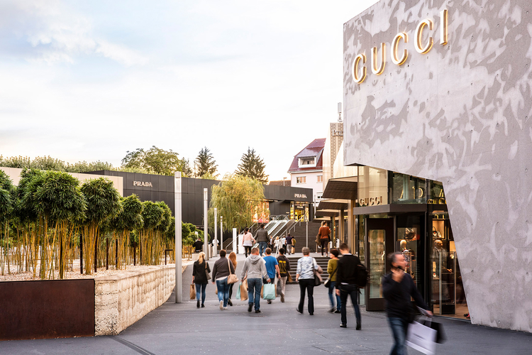 Die Szene zeigt eine belebte Einkaufsstraße mit Menschen, die zwischen modernen Geschäften flanieren. Auf der rechten Seite ist das Geschäft von Gucci sichtbar, während ein Teil von Prada im Hintergrund zu sehen ist. Grüne Pflanzen rahmen den Fußweg ein.