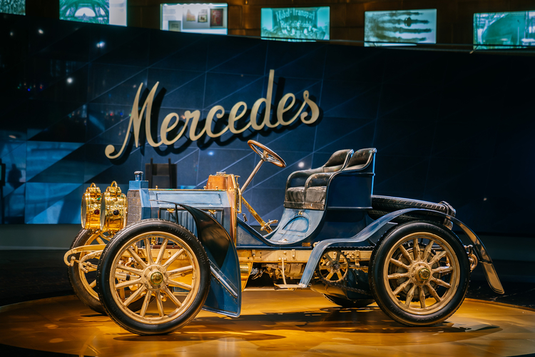 Ein historisches Auto der Marke Mercedes steht im Vordergrund. Es hat eine blaue Karosserie, goldene Details und große, speichenräder. Im Hintergrund ist der Schriftzug „Mercedes“ an einer Wand zu sehen. Die Umgebung ist modern gestaltet, was den Kontrast zur klassischen Fahrzeugform verstärkt.
