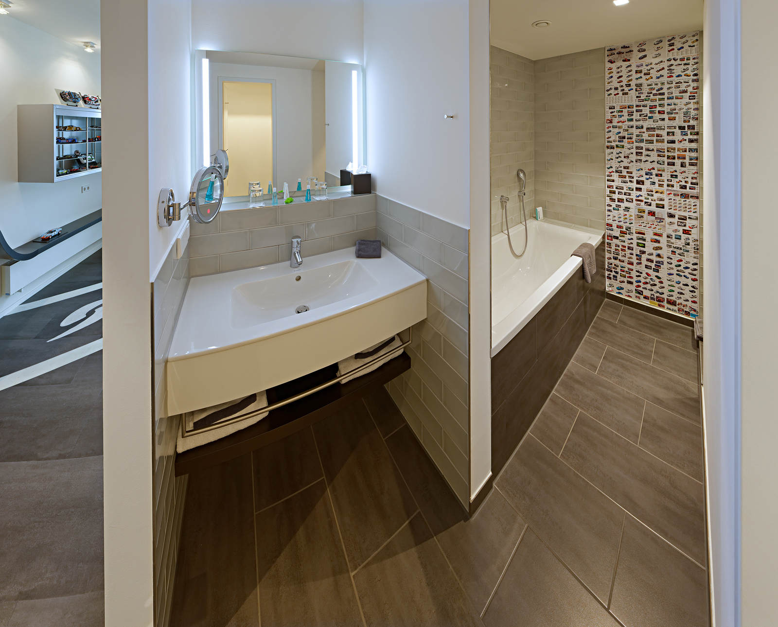 Ein modernes Badezimmer mit einem Waschbecken, Spiegel und einer Badewanne. Der Boden ist mit großen, grauen Fliesen ausgestattet. An einer Wand sind bunte Bilder angebracht. Die Beleuchtung ist hell und schafft eine einladende Atmosphäre.