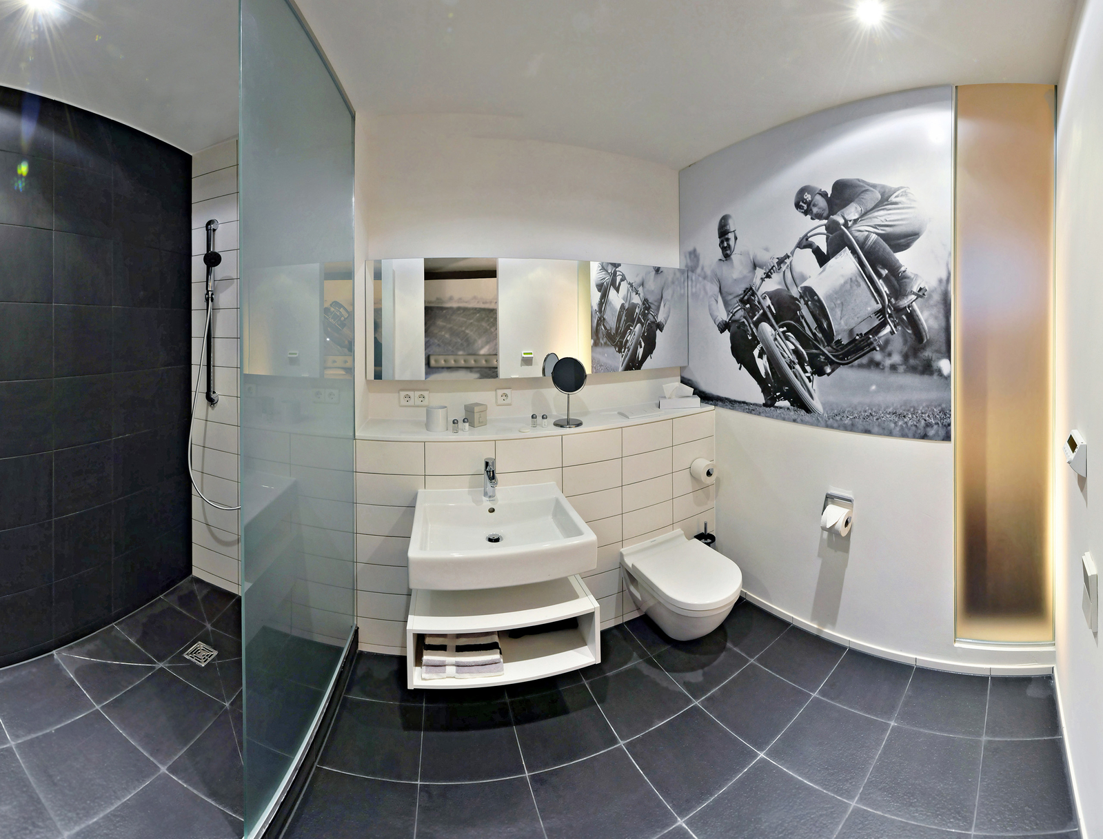 Das Bild zeigt ein modernes Badezimmer mit einer großen Dusche, einem Waschbecken und einem WC. An der Wand befindet sich eine Schwarz-Weiß-Fotografie, die zwei Männer auf Motorrädern darstellt. Der Boden ist dunkel gefliest und die Wände sind hell gestaltet.