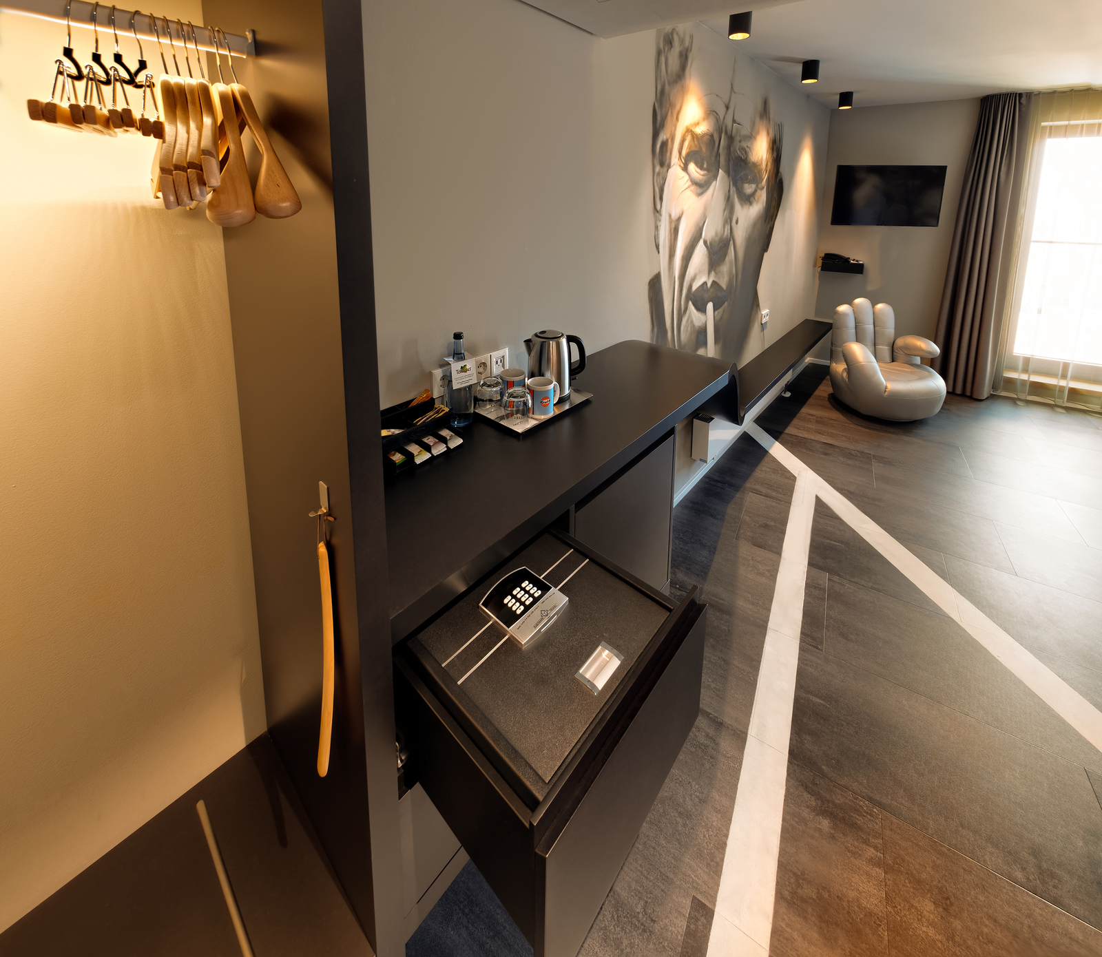 Das Bild zeigt eine moderne Hotelzimmer-Einrichtung mit einem schwarzen Tisch, einem integrierten Safe mit Zahlenkombination, einem Wasserkocher und Getränken. An der Wand sind hölzerne Kleiderbügel sowie ein beeindruckendes Wandbild einer Person zu sehen. Das Zimmer hat große Fenster, die Tageslicht hereinlassen.