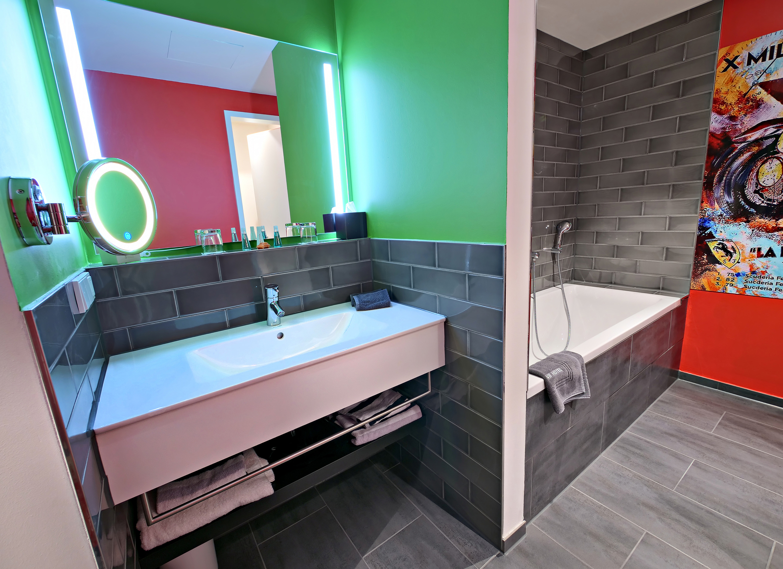 Das Bild zeigt ein modernes Badezimmer mit einem Waschbecken, einem großen Spiegel und einer Badewanne. Die Wände sind in lebendigen Farben gestrichen: Grün und Rot. Am Waschbecken befinden sich Handtücher und Pflegeartikel. Der Raum hat stilvolle graue Fliesen und eine praktische Beleuchtung.