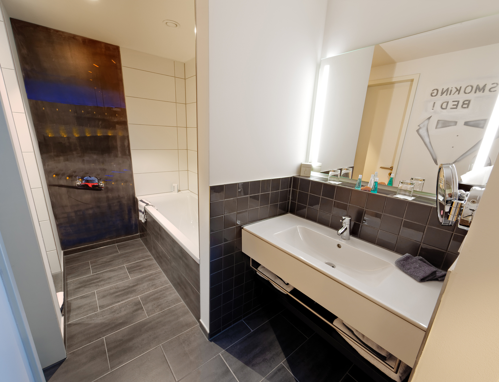Ein modernes Badezimmer mit einer großen Badewanne auf der linken Seite und einem Waschbecken auf der rechten Seite. Der Boden ist mit dunklen Fliesen ausgelegt. An der Wand sind helle Fliesen und ein Spiegel mit Beleuchtung angebracht. Im Hintergrund ist ein dekorativer Akzent zu sehen.