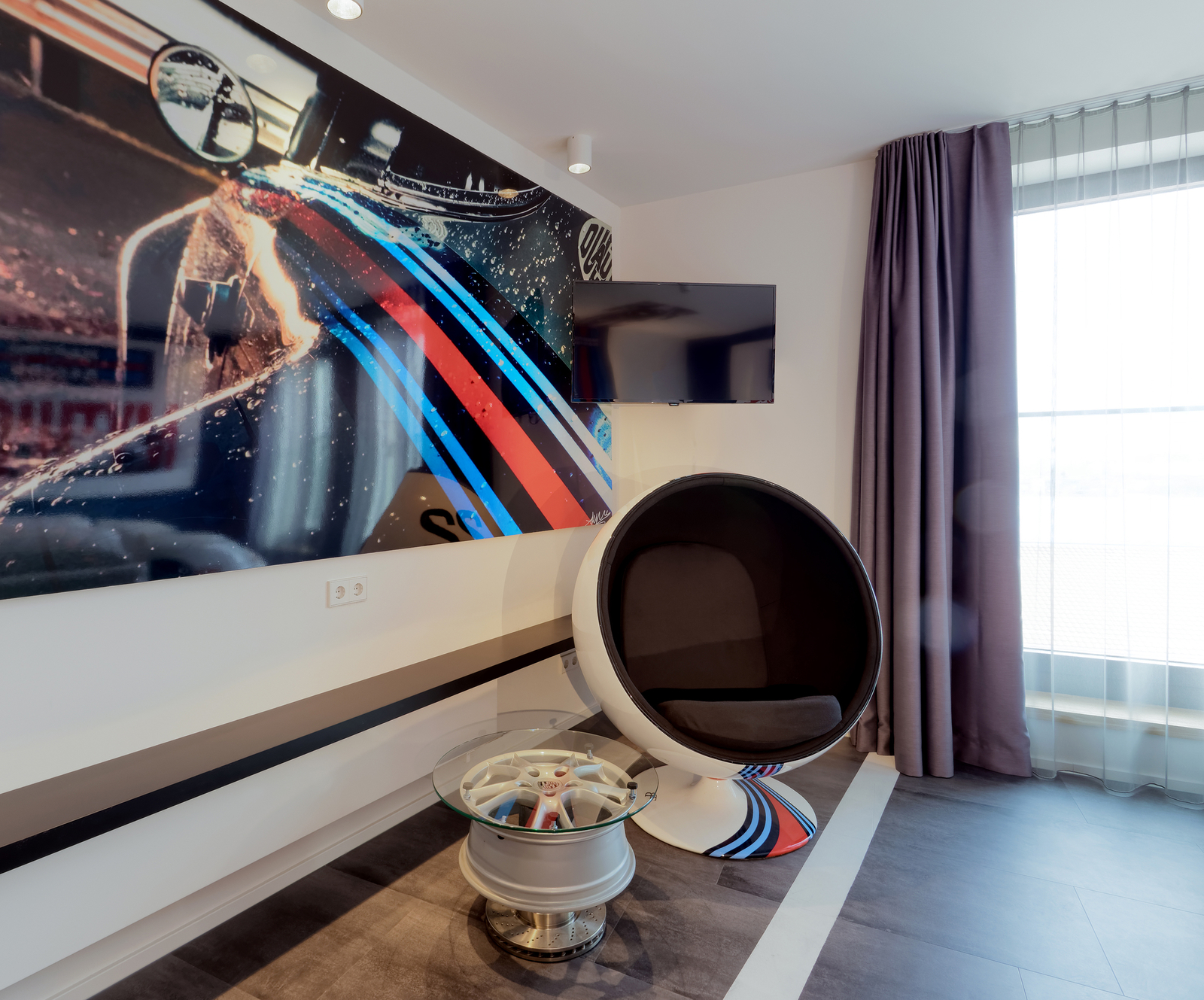 Das Bild zeigt einen modernen Raum mit einem runden Sessel, der auf einem auffälligen Sockel steht. Ein Glascouchtisch mit einem Rad als Tischunterlage ist daneben. Im Hintergrund hängt ein großes Wandbild eines Autos mit bunten Streifen. Ein Fernseher ist an der Wand montiert.