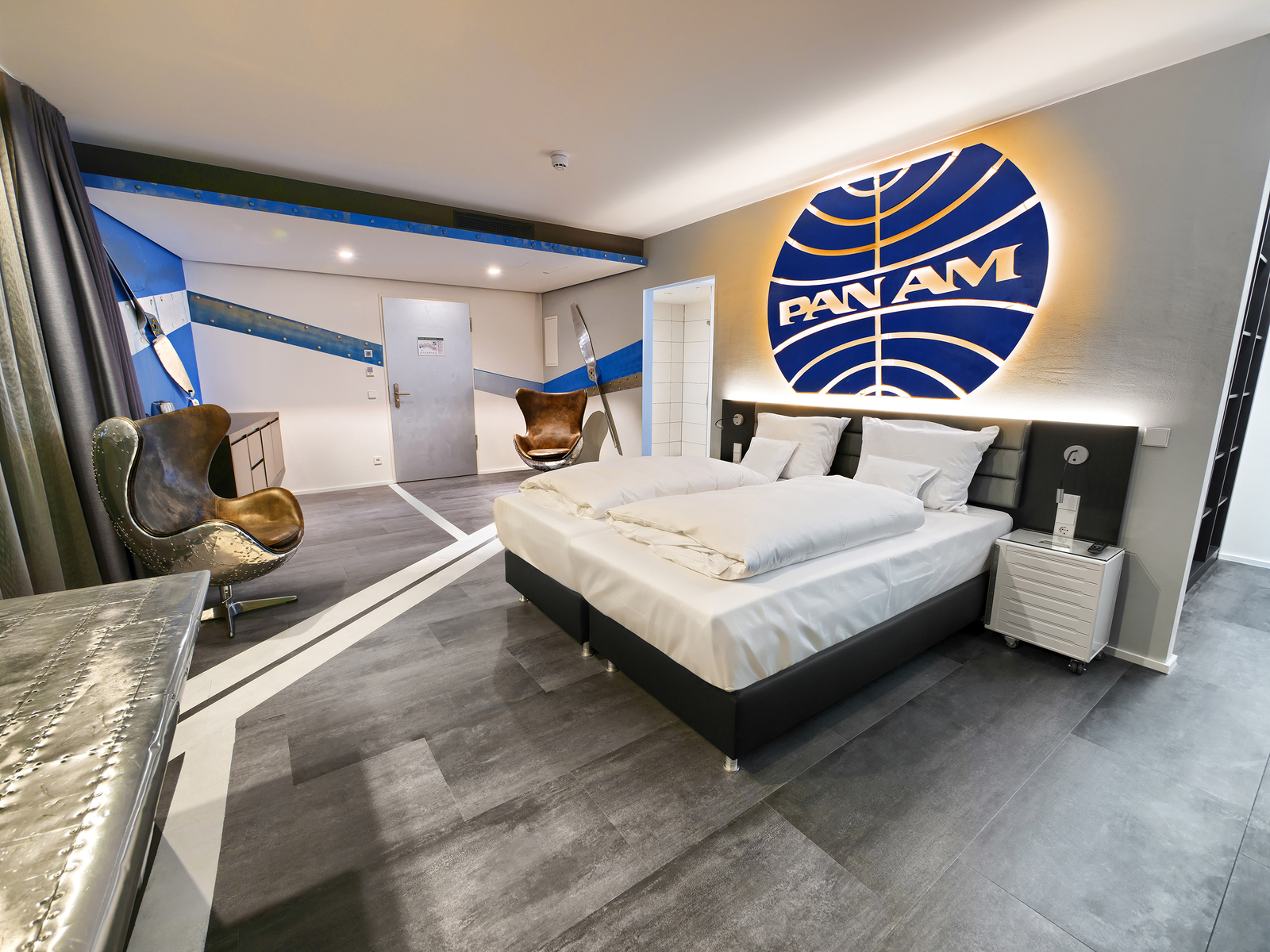Großzügiges Doppelbett unter einem leuchtenden PanAm Logo gegenüber eines Ledersessels im Themenzimmer des V8 Hotels