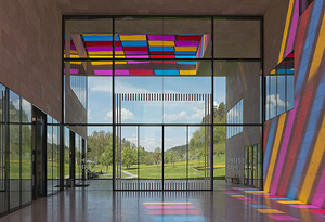 Ein heller Innenraum eines Museums mit großen Fensterfronten. Bunte Lichtstrahlen in Rosa, Gelb und Blau fallen auf den Boden. Draußen sind grüne Wiesen und Bäume sichtbar. Der Raum vermittelt eine offene und einladende Atmosphäre.