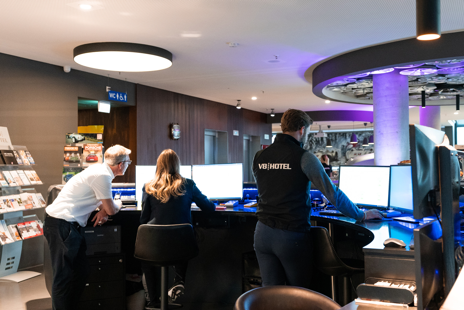 Im Bild sind drei Personen in einem modernen Empfangsbereich zu sehen. Zwei sitzen an Schreibtischen mit mehreren Computermonitoren, während eine Person mit dem Rücken zur Kamera steht und einen Monitor bedient. Der Raum hat eine elegante, zeitgenössische Gestaltung.
