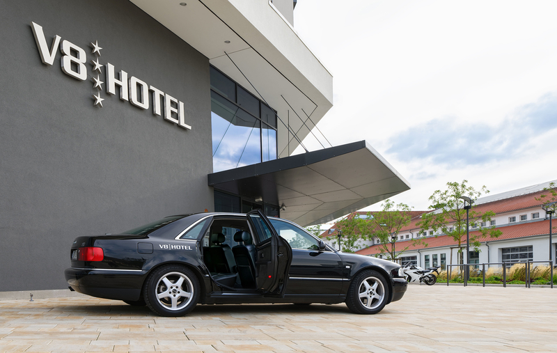 Ein schwarzes Auto mit offener Tür steht vor dem V8 Hotel. Die Fassade des Hotels trägt den Schriftzug V8 HOTEL in großen, modernen Buchstaben. Im Hintergrund sind Bäume und andere Gebäude sichtbar. Der Himmel ist teilweise bewölkt.