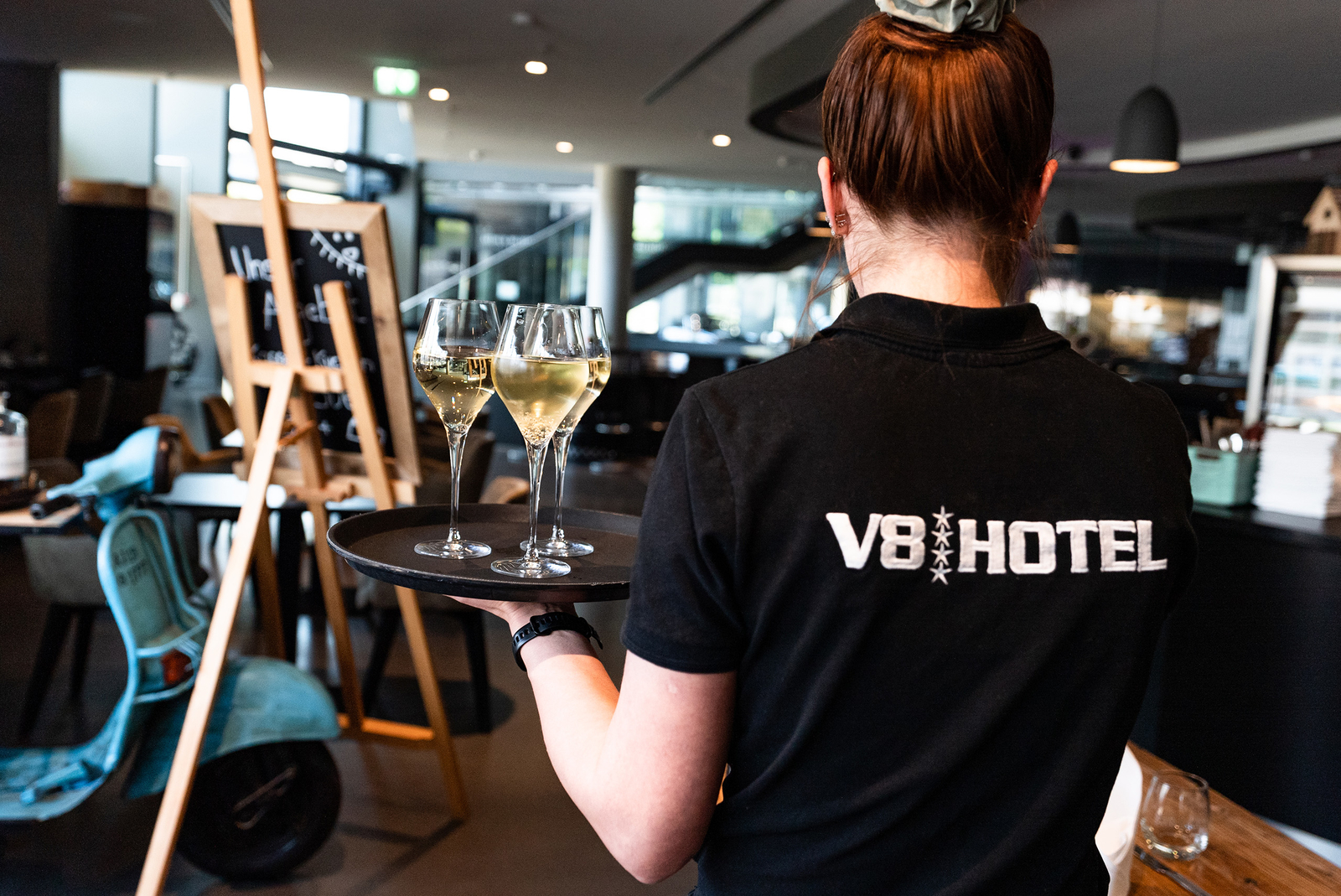 Eine Kellnerin trägt ein Tablett mit zwei Gläsern Sekt. Sie steht in einem modernen Restaurant mit stilvollem Ambiente. Im Hintergrund ist eine Tafel zu sehen, die Informationen anzeigt.