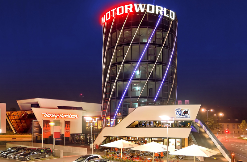 Das Bild zeigt das Gebäude von MOTORWORLD bei Nacht. Es hat eine moderne, gläserne Architektur mit illuminierter Beschriftung. Im Vordergrund ist ein Restaurantbereich mit Sonnenschirmen sichtbar, daneben steht ein Harley-Davidson-Shop. Die Umgebung ist urban mit wenigen Autos auf dem Parkplatz.