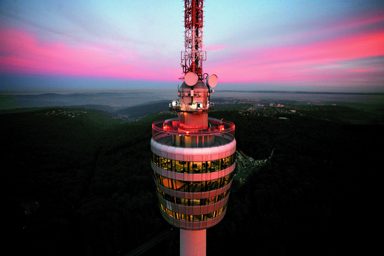 Ein hoher Aussichtsturm mit einer runden Plattform und großen Fenstern, von der ein Blick ins weite Tal geboten wird. Der Himmel ist in sanften Rosa- und Blautönen gefärbt, während die Stadt und Wälder im Hintergrund sichtbar sind. Die Plattform ist schwach beleuchtet.