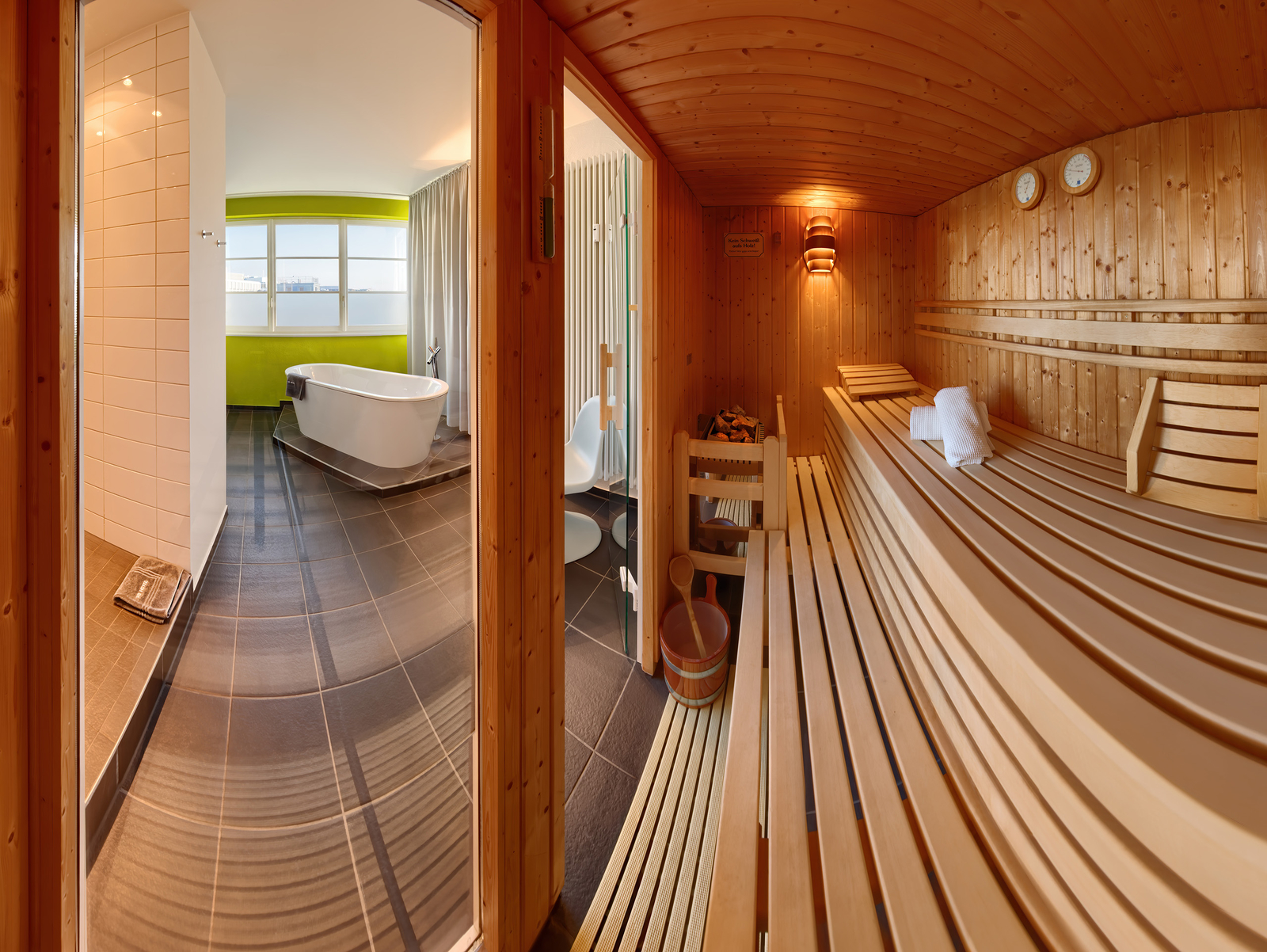 Ein Blick in eine helle Sauna aus Holz, mit mehreren Holzliegen und Handtüchern. Durch die Glastür sieht man einen modernen Wellnessbereich mit einer Badewanne und großen Fenstern, die viel Tageslicht hereinlassen. Die Atmosphäre ist entspannend und einladend.