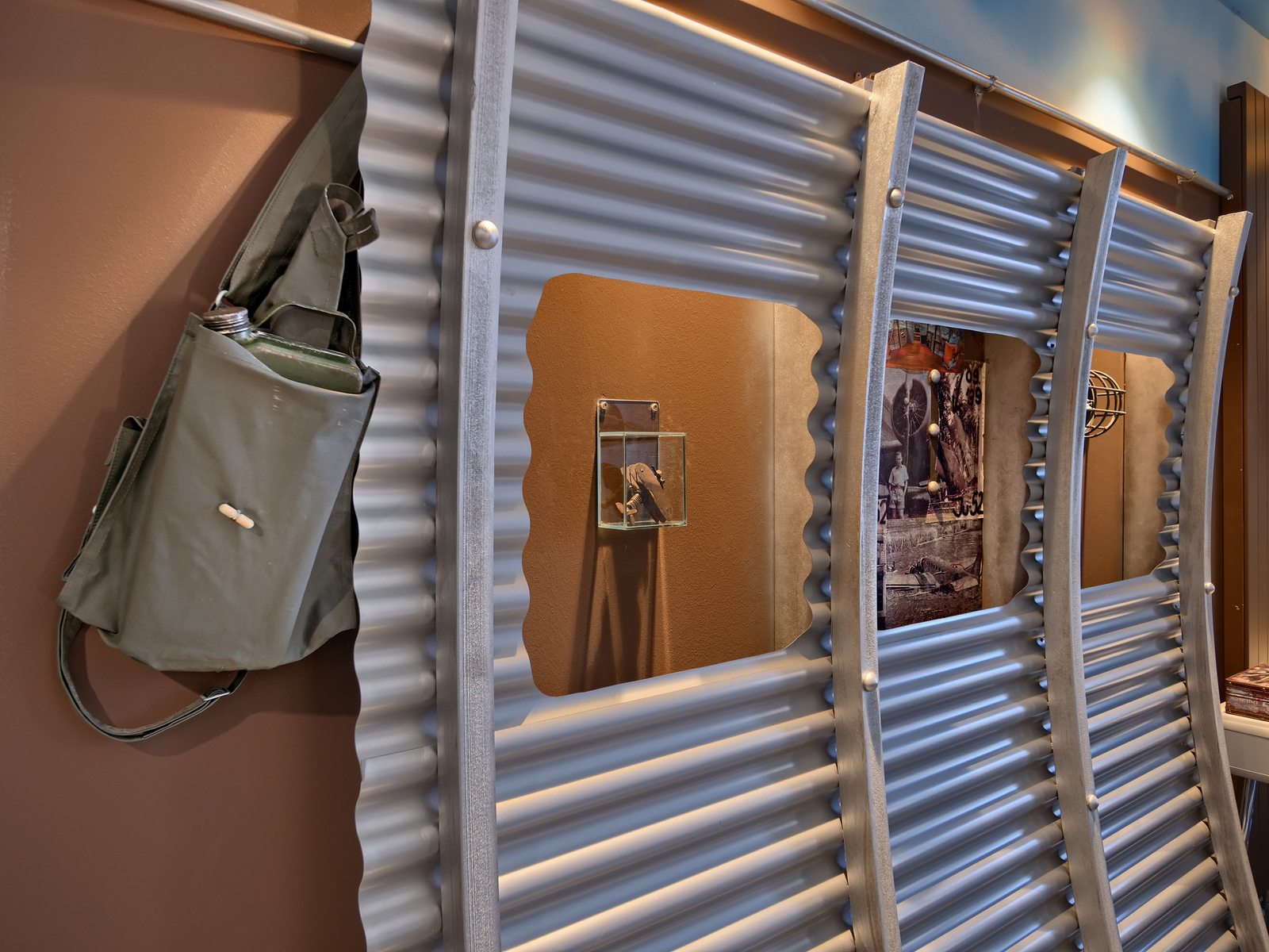 Ein graues, gewelltes Metallpaneel bildet eine Wand. An der Wand hängt eine grüne Tasche mit einer Wasserflasche. In einer Nische ist ein kleines Glasdisplay, das ein Objekt zeigt. Im Hintergrund ist ein Wandbild, das eine historische Szene darstellt.