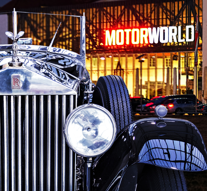 Ein klassischer Rolls-Royce mit glänzendem Chrom und einem markanten Kühlergrill ist im Vordergrund zu sehen. Im Hintergrund leuchtet das Schild MOTORWORLD an einer modernen Gebäude-Fassade, die eine automobile Erlebniswelt symbolisiert.