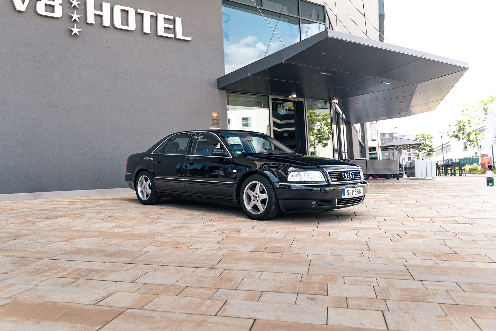 Ein schwarzer Audi steht vor dem V8 Hotel. Das Fahrzeug ist auf einem gepflasterten Bereich abgestellt, hinter dem sich die moderne Fassade des Hotels befindet. Am Eingang sind große Fenster sichtbar, und einige Pflanzen sind im Hintergrund angeordnet.