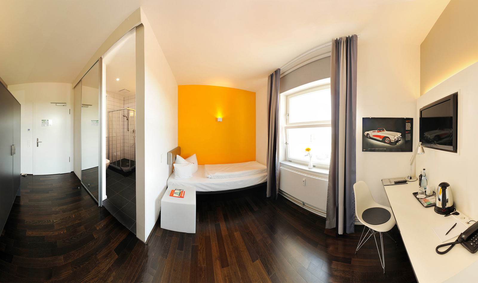 Die Abbildung zeigt ein helles Hotelzimmer mit einem Einzelbett, einem Arbeitsplatz und einem Fenster mit Blick nach draußen. Die Wände sind in warmen Farben gehalten, und es gibt einen kleinen Tisch neben dem Bett. Im Badezimmerbereich ist eine Dusche sichtbar.