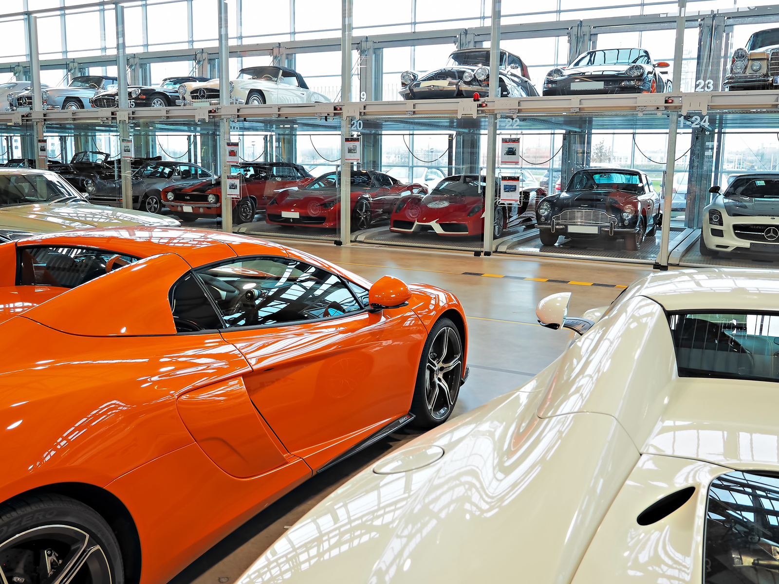 In der Motorworld stehen ein orangefarbener Sportwagen und ein weißes Fahrzeug im Vordergrund. Im Hintergrund sind mehrere weitere Oldtimer und Sportwagen in regalförmigen Vitrinen angeordnet, die gut beleuchtet sind.