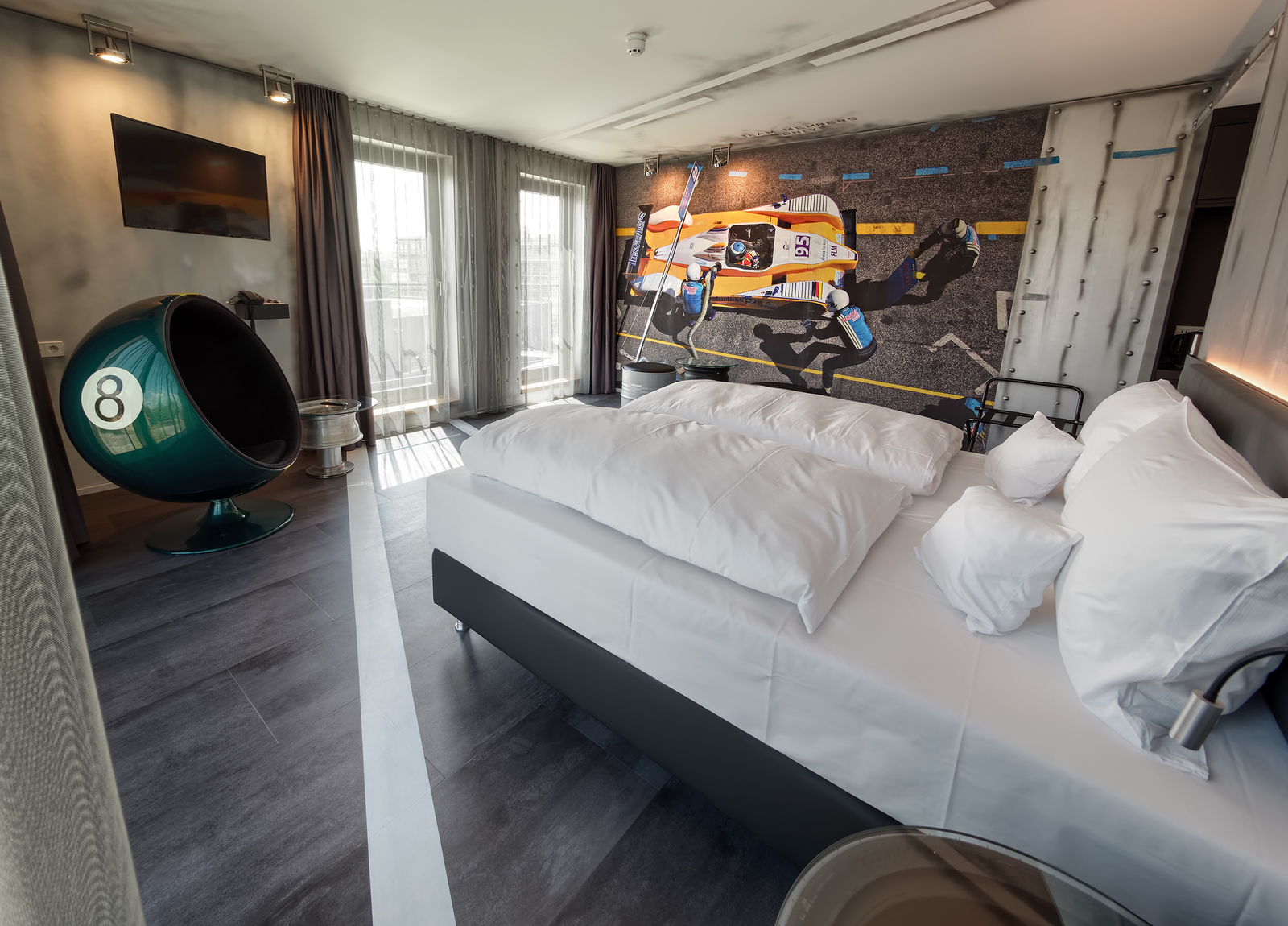 Ein modernes Schlafzimmer mit einem großen Doppelbett, das mit weißen Kissen und Decken ausgestattet ist. An der Wand befindet sich ein auffälliges Wandbild eines Rennwagens. In der Ecke steht ein stilvoller grüner Sessel mit der Nummer 8. Fenster lassen viel Licht herein.