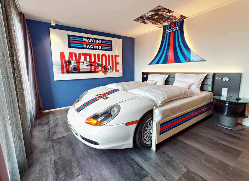 Das Bild zeigt ein stylisches Schlafzimmer, das mit einem Bett im Design eines Rennwagens ausgestattet ist. Das Bett hat einen weißen Aufsatz mit einer bunten „Martini Racing“-Lackierung. Im Hintergrund ist ein großes Wandbild mit der Aufschrift „MYTHIQUE“ und einem Rennwagenmotiv zu sehen.
