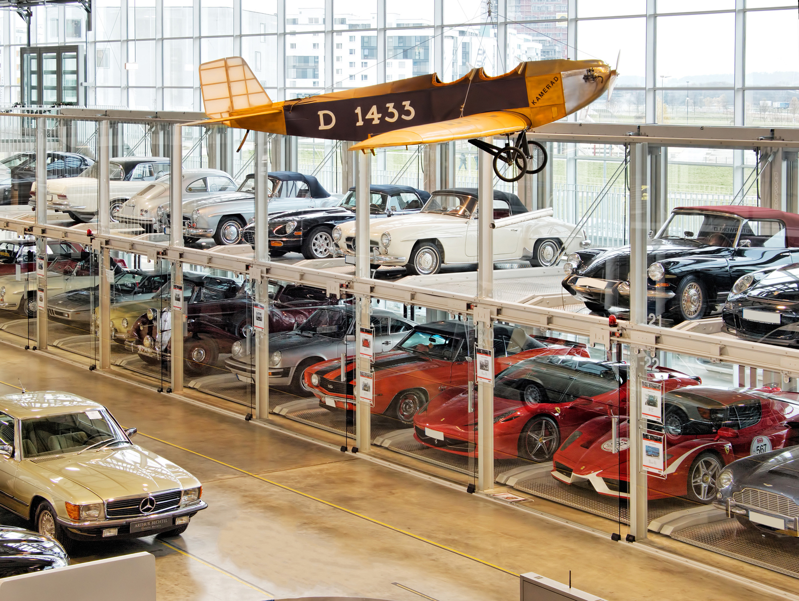 Das Bild zeigt eine moderne Autogarage mit mehreren klassischen Autos, die hinter Glasabtrennungen ausgestellt sind. An der Decke hängt ein historisches Flugzeug. Die Umgebung wirkt hell und offen, mit großen Fenstern, die den Blick nach draußen ermöglichen.