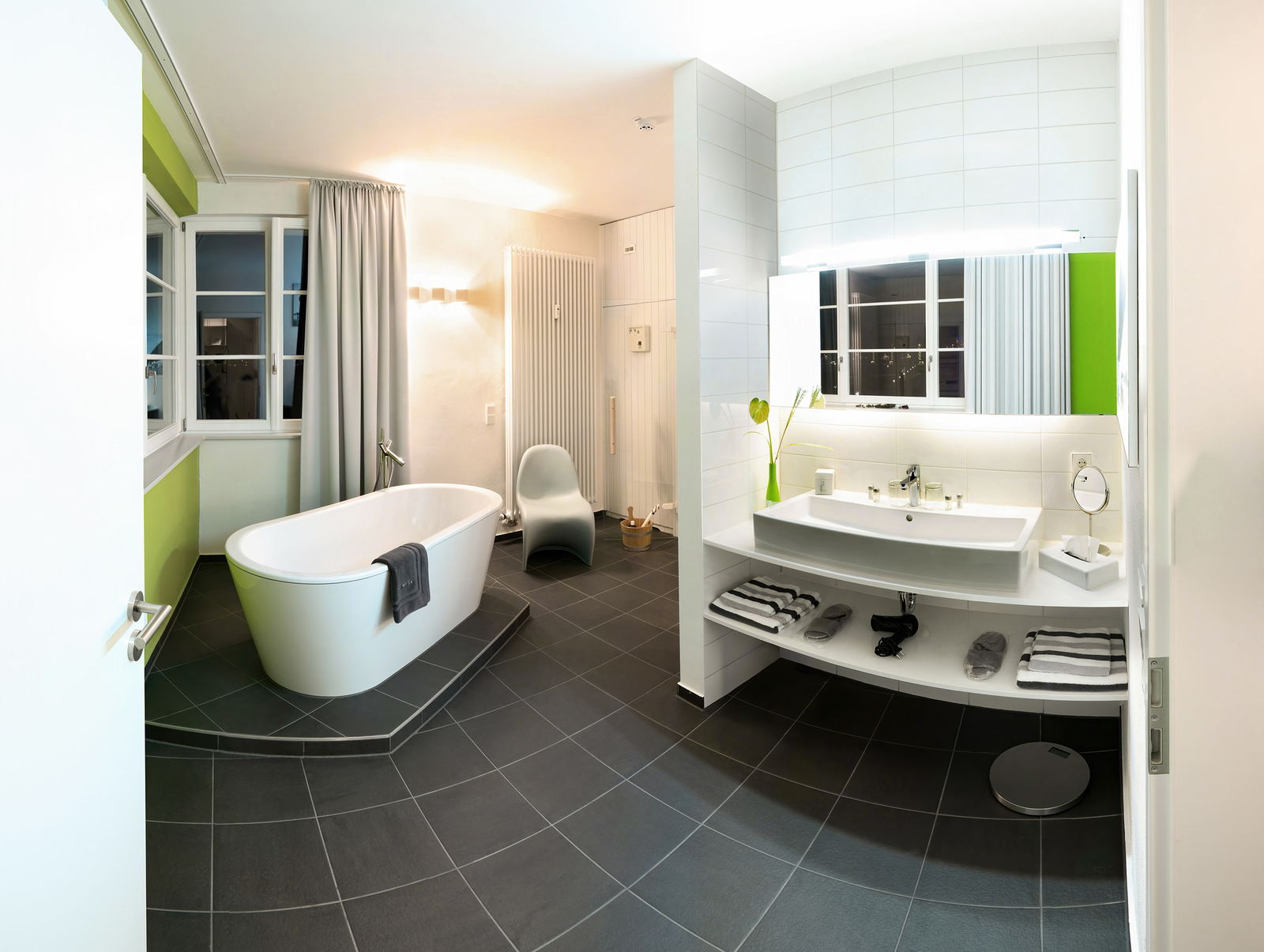 Ein modernes Badezimmer mit freistehender Badewanne, einem eleganten Waschbecken und einem großen Spiegel. Die Wände sind in hellen Farben mit einem grünen Akzent gestaltet. Handtücher liegen ordentlich auf einem Regal, und große Fenster sorgen für viel Tageslicht.