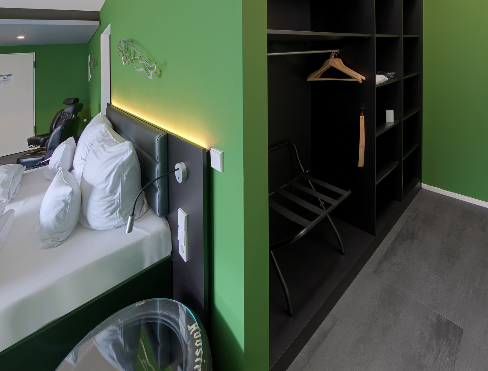 Das Bild zeigt ein modernes Zimmer mit grünen Wänden. Auf der linken Seite sind ein Bett mit weißen Kissen und eine Leselampe zu sehen. Rechts befindet sich ein begehbarer Kleiderschrank mit einem Haken und einem Stuhl. Der Boden ist grau gefliest.