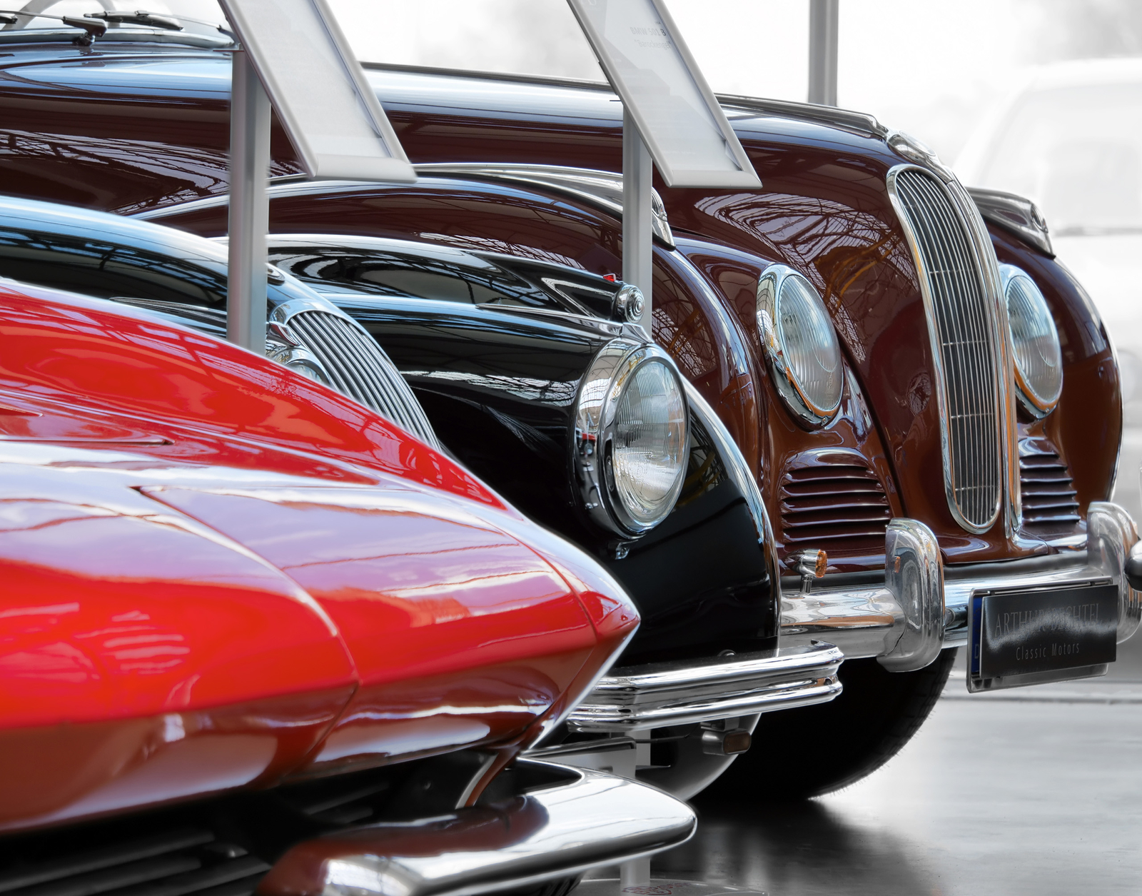 Eine Reihe klassischer Autos sind in einer Ausstellung aufgereiht. Im Vordergrund ist ein rotes Auto sichtbar, gefolgt von einem schwarzen und einem braunen Fahrzeug. Jedes Auto zeigt markante Scheinwerfer und verchromte Details, die ihre Eleganz und historischen Wert betonen.