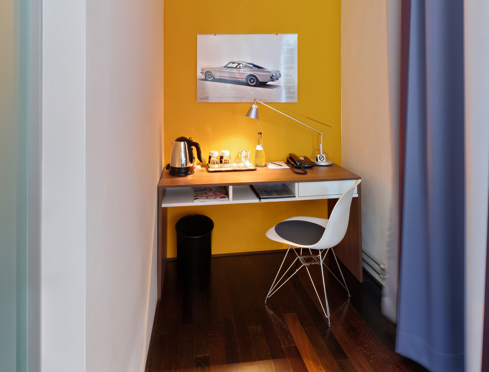 Ein kleiner Arbeitsplatz in einer Ecke eines Raumes. Eine Holzplatte mit einer Lampe, einem Wasserkrug, Gläsern und einem Telefon. An der Wand hängt ein Bild eines Autos auf orangefarbenem Hintergrund. Der Boden ist aus dunklem Holz.