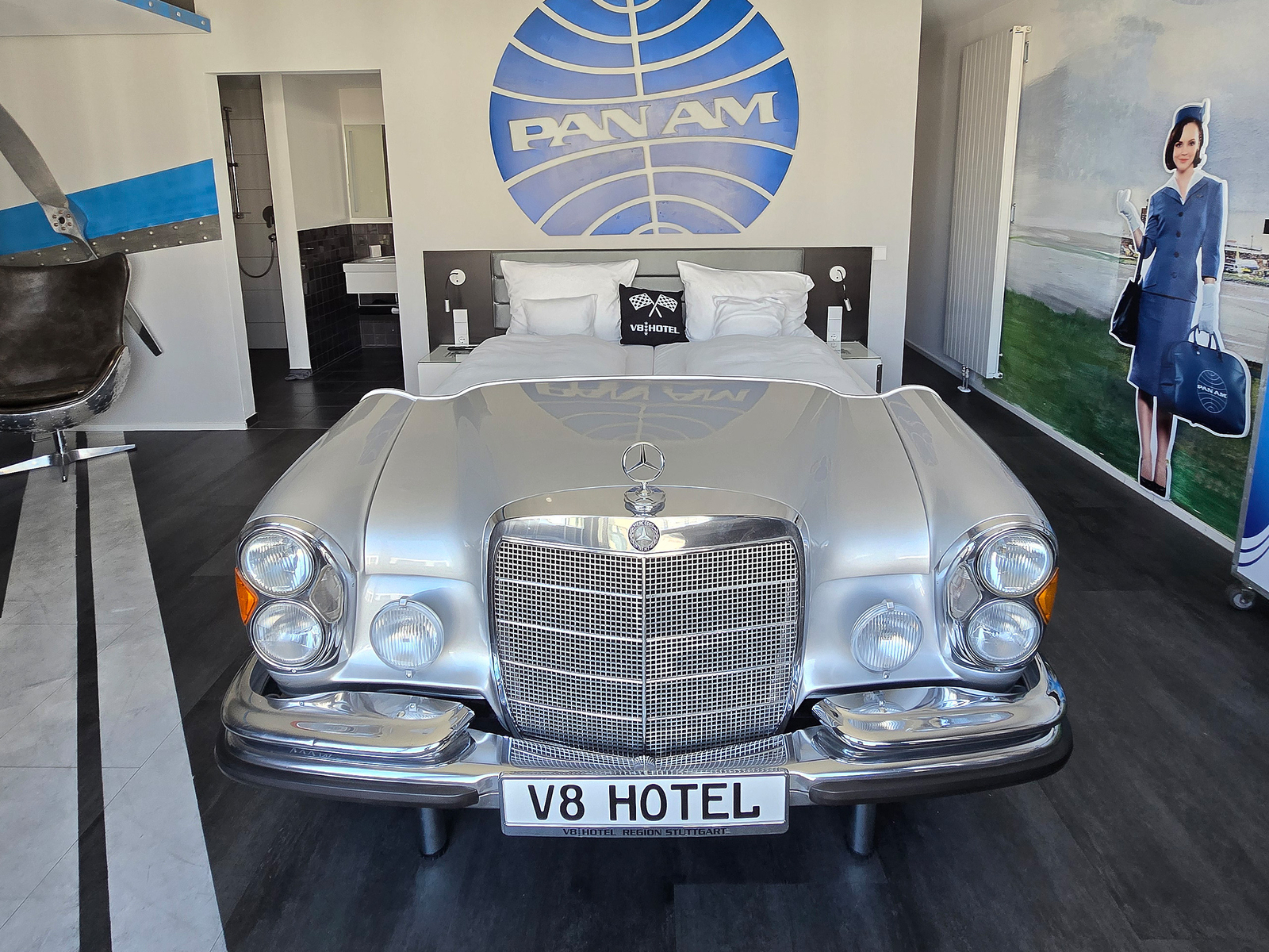 Ein silberner Mercedes-Benz mit Frontansicht dient als Bett in einem modernen Hotelzimmer. Im Hintergrund ist ein Pan Am-Logo und eine lebensgroße Figur einer Stewardess zu sehen, die an eine Flugreise erinnert. Das Zimmer hat ein stilvolles, zeitgemäßes Design.