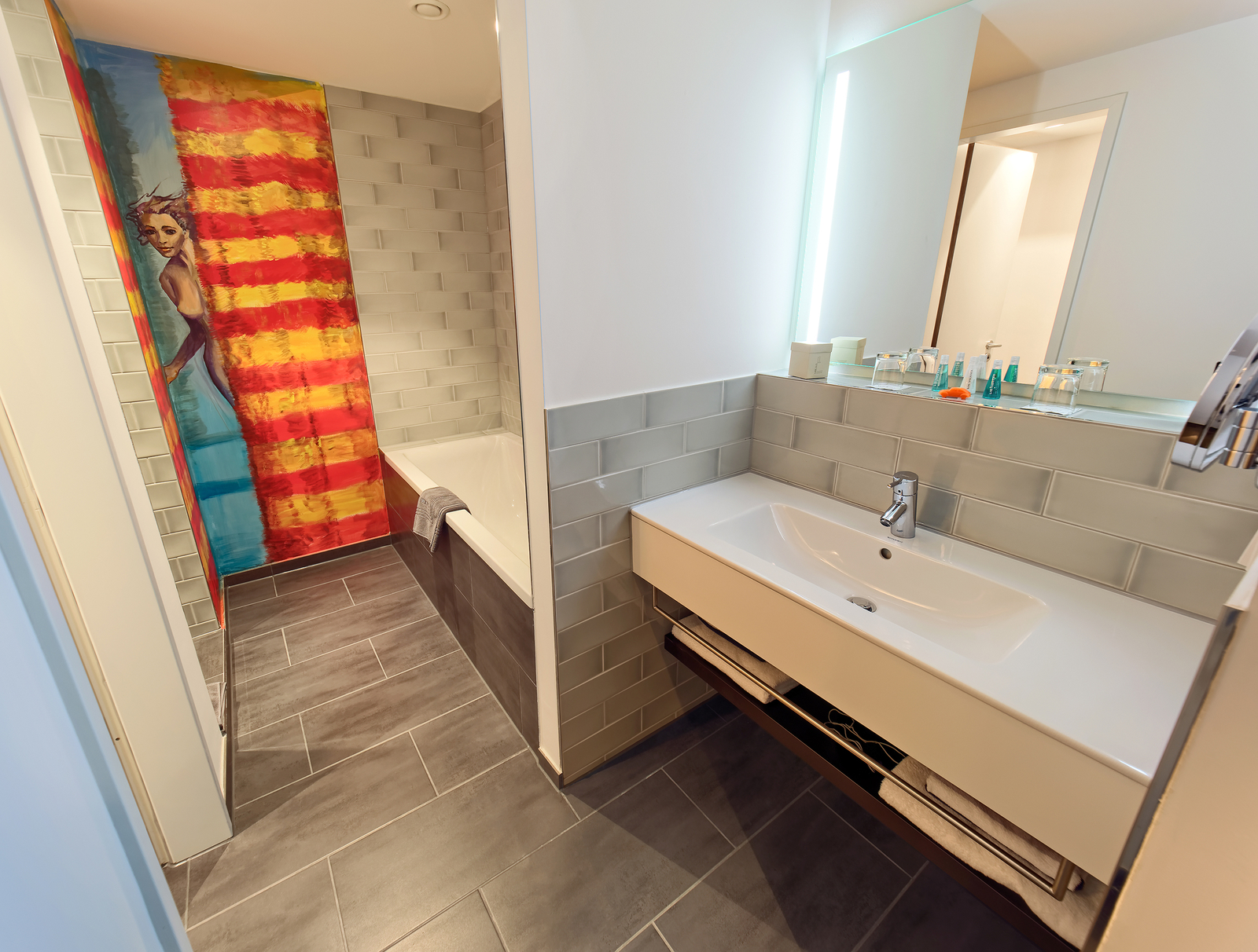 Das Bild zeigt ein modernes Badezimmer mit einer schlanken Waschtischplatte, einem großen Spiegel und einer Badewanne. An einer Wand befindet sich ein buntes Wandgemälde mit einer stilisierten Darstellung, das einen lebhaften Farbakzent setzt. Der Boden ist mit grauen Fliesen ausgelegt.